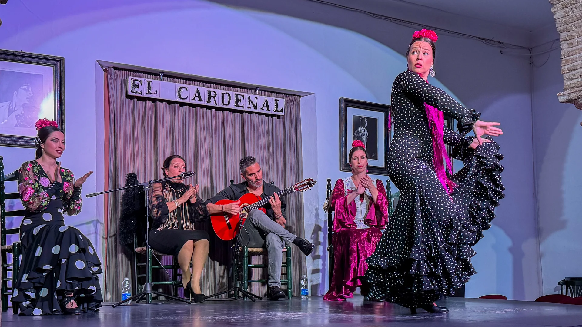 Flamenková tanečnice v černých puntíkatých šatech uprostřed otáčky na jevišti Tablao El Cardenal, za ní kytarista, zpěvák a dvě sedící tanečnice pod obloženým nápisem s názvem